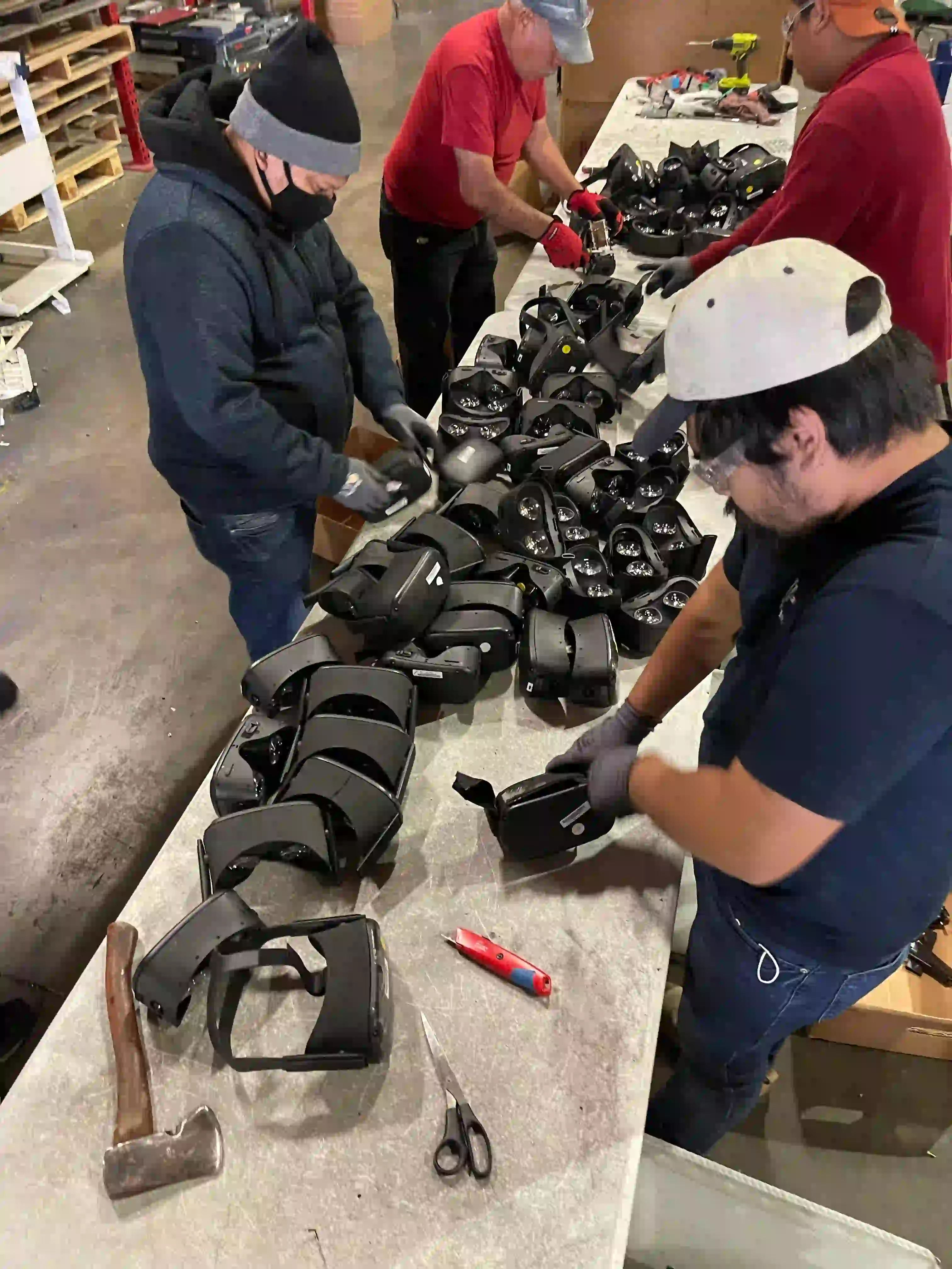 Workers assembling and inspecting various black products on a table, surrounded by tools like scissors and an axe.