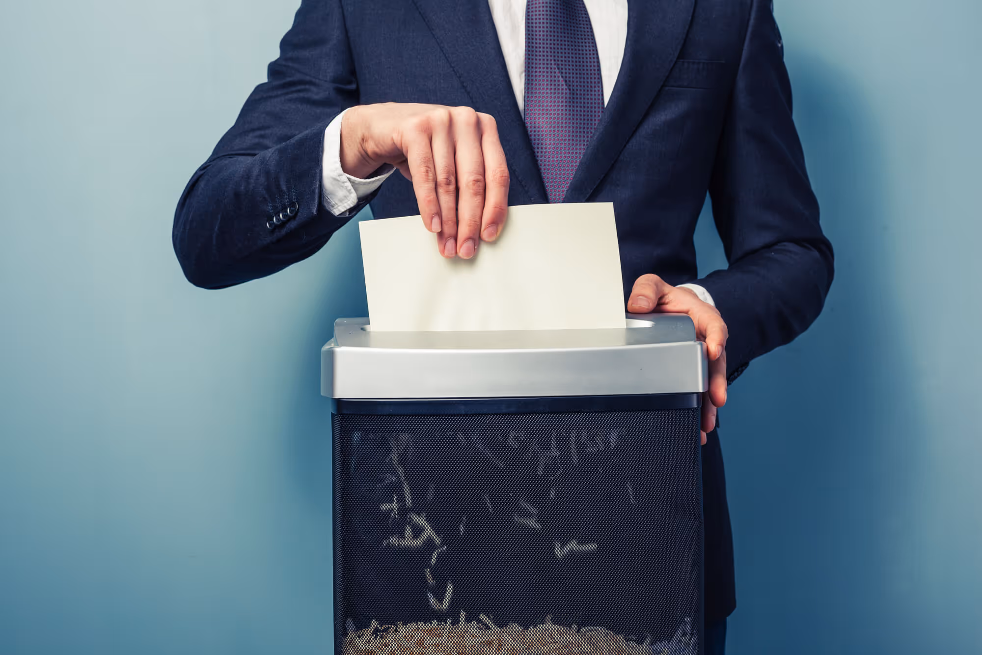 A person in a dark suit and tie feeds a blank white sheet of paper into a paper shredder