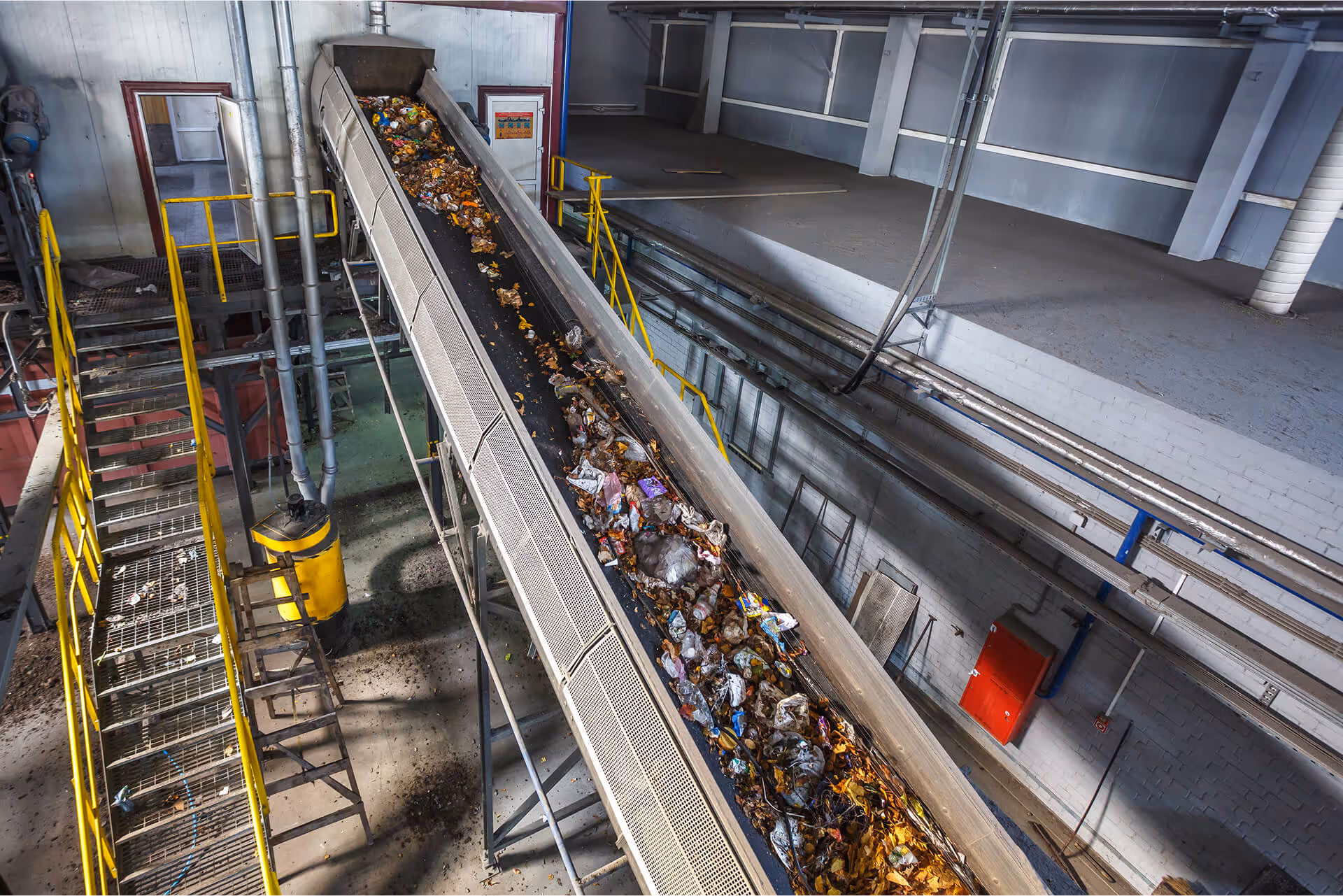  Overhead view of a long conveyor belt carrying a mix of trash and waste material inside a recycling or waste sorting facility, next to yellow stairs