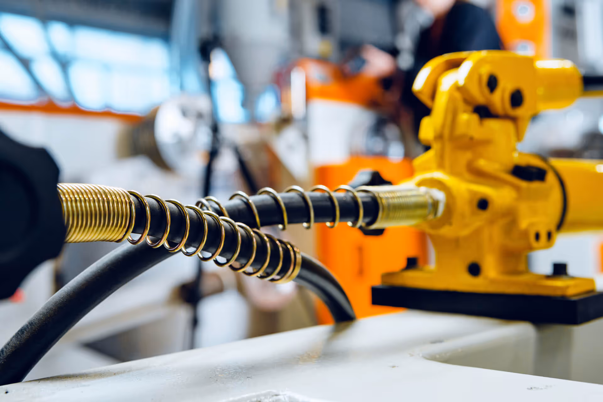 Close-up on two black industrial hoses wrapped in golden spring coils, connecting to a bright yellow machine in a blurred factory background