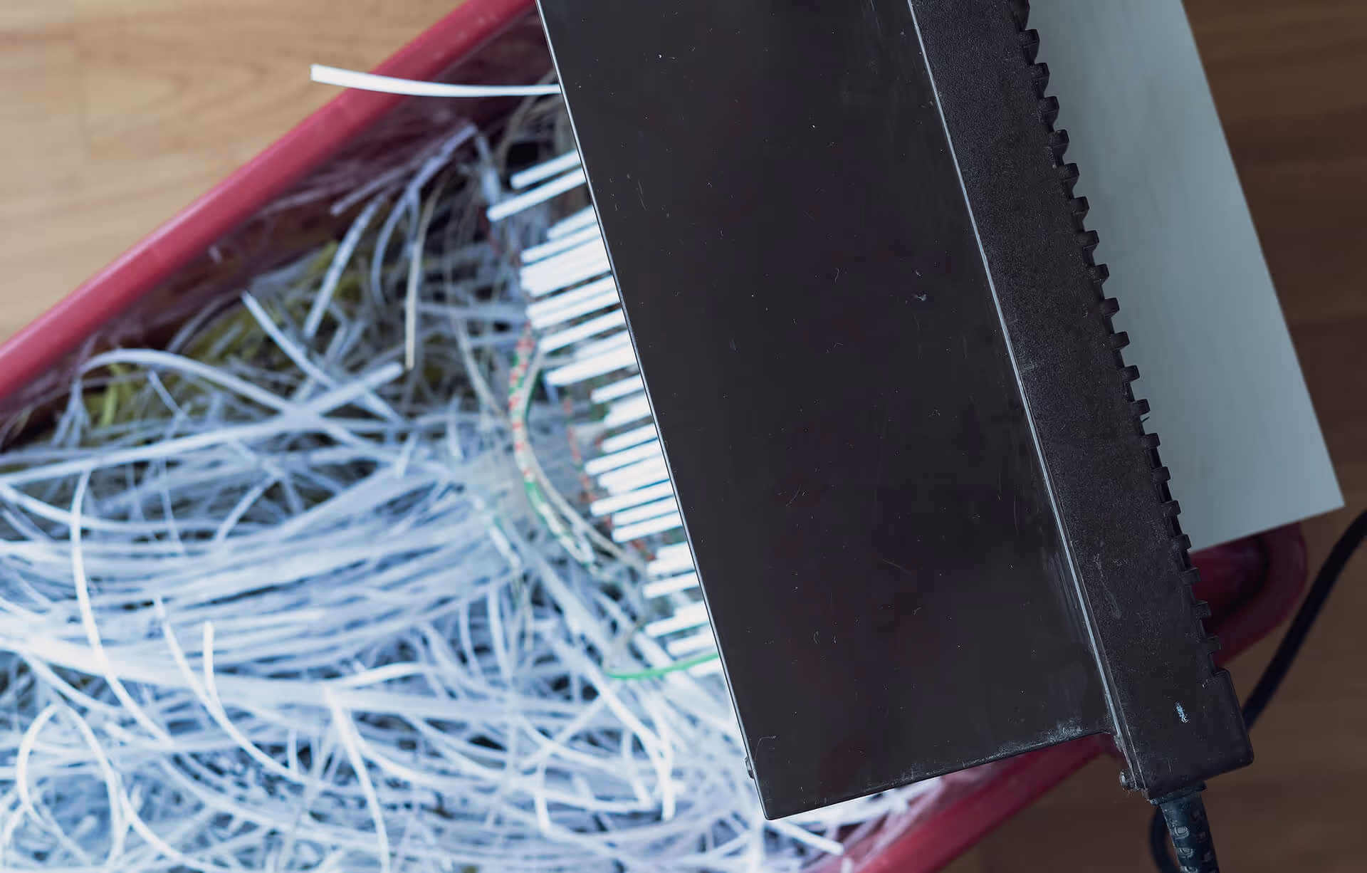 Close-up of a paper shredder's black head resting on a red bin filled with long, thin strips of shredded white paper.