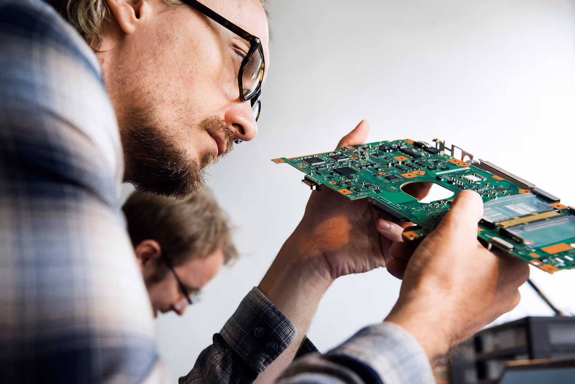 A close-up side view of a technician wearing glasses intently inspecting a green computer motherboard