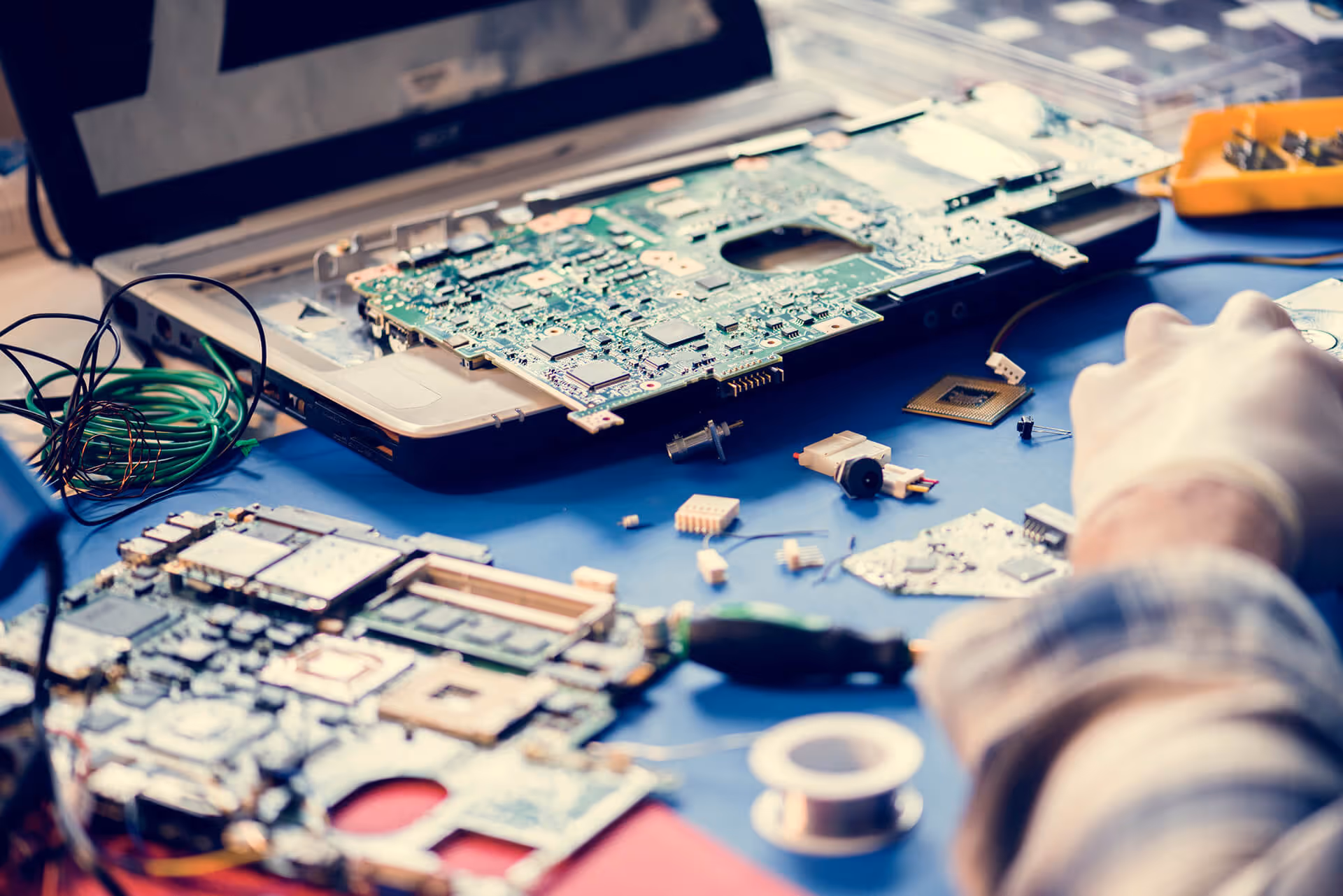 A technician repairs a laptop motherboard on a workspace filled with electronic components, microchips, and soldering tools