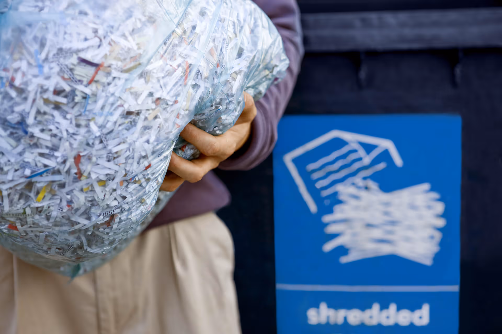 Person holding a large bag of shredded paper next to a blue recycling bin