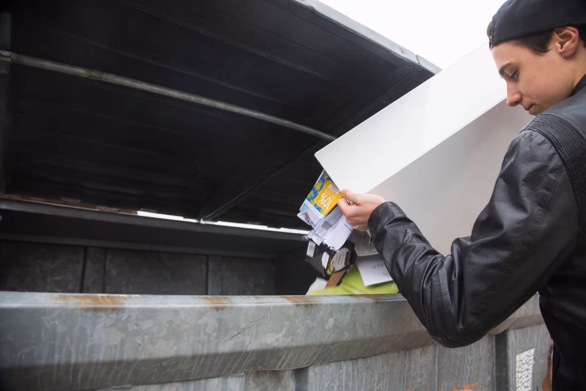Young man emptying a container of paper waste into a large recycling dumpster