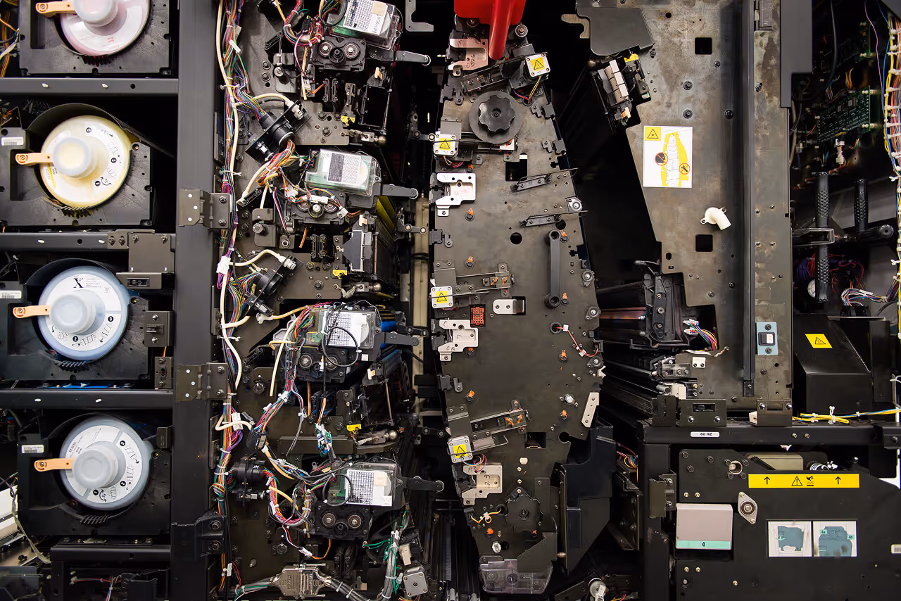 Internal view of a professional high-volume printer showing complex mechanical gears, wiring, and toner cartridges